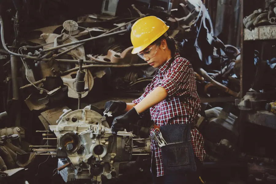 La Reducción de la Jornada Laboral: Evolución, Beneficios y Desafíos Actuales 3 Una trabajadora con casco de seguridad amarillo y gafas protectoras ajusta una máquina en un taller industrial. Viste una camisa de cuadros y un cinturón de herramientas, demostrando su destreza y compromiso con el trabajo. El entorno está lleno de maquinaria y piezas mecánicas, resaltando el ambiente de un taller en pleno funcionamiento.